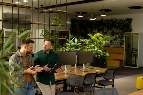 Two professionals are engaged in a focused conversation while holding digital tablet in a contemporary office space, featuring rustic exposed brickwork and warm natural light - Photo, Image