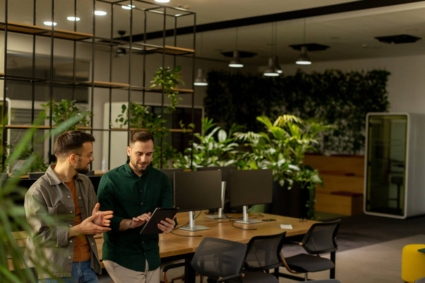 Два профессионала ведут целенаправленную беседу, держа в руках цифровой планшет в современном офисном помещении, с элементами деревенского кирпича и теплым естественным светом - Фото, изображение