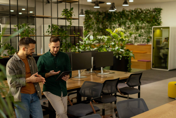Dwóch profesjonalistów zajmuje się skoncentrowaną rozmową trzymając tablet cyfrowy we współczesnej przestrzeni biurowej, wyposażony w rustykalne odsłonięte cegły i ciepłe naturalne światło - Zdjęcie, obraz