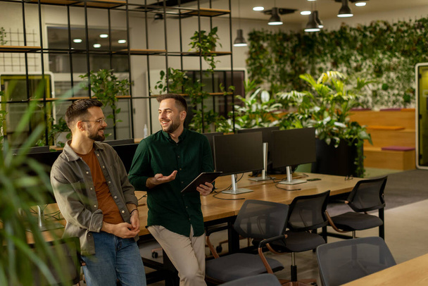 Dos profesionales se dedican a una conversación enfocada mientras sostienen la tableta digital en un espacio de oficina contemporáneo, con ladrillos expuestos rústicos y luz natural cálida - Foto, Imagen