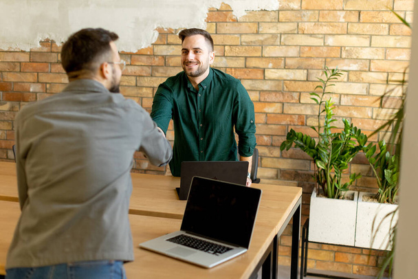 Two professionals engage in a welcoming handshake across a wooden table adorned with laptops, signaling a successful meeting or partnership in a contemporary brick-walled office - Фото, изображение