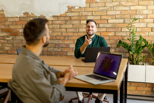 Two smiling professionals engage in a collaborative work session at a wooden table, their camaraderie evident in a contemporary office setting with an exposed brick wall backdrop - Photo, Image