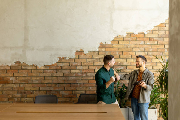 Deux professionnels s'engagent dans une conversation ciblée tout en tenant une tablette numérique dans un espace de bureau contemporain, mettant en vedette des briques rustiques exposées et une lumière naturelle chaude - Photo, image