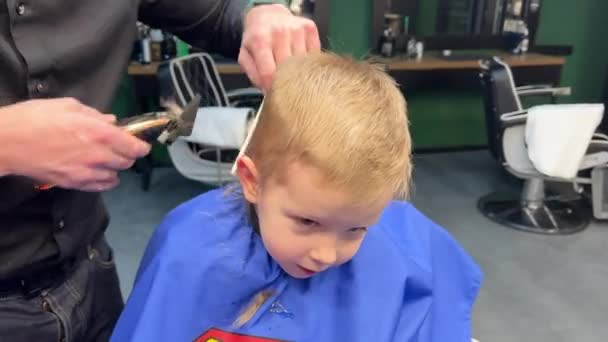 Kappers gebruikt elektrisch scheerapparaat om jongens haar te knippen. Het kleine kind krijgt het eerste kapsel in de kapperszaak. Sluiten. - Video