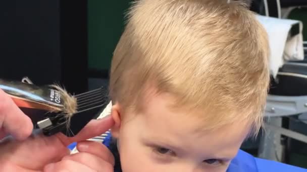 Kappers gebruikt elektrisch scheerapparaat om jongens haar te knippen. Het kleine kind krijgt het eerste kapsel in de kapperszaak. Sluiten. - Video