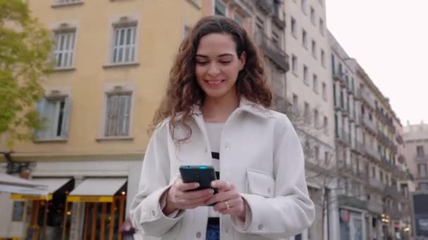Mooie jonge vrouw met behulp van mobiele telefoon staan in de stad straat. Gelukkig glimlachende vrouw chatten op smartphone app. - Video