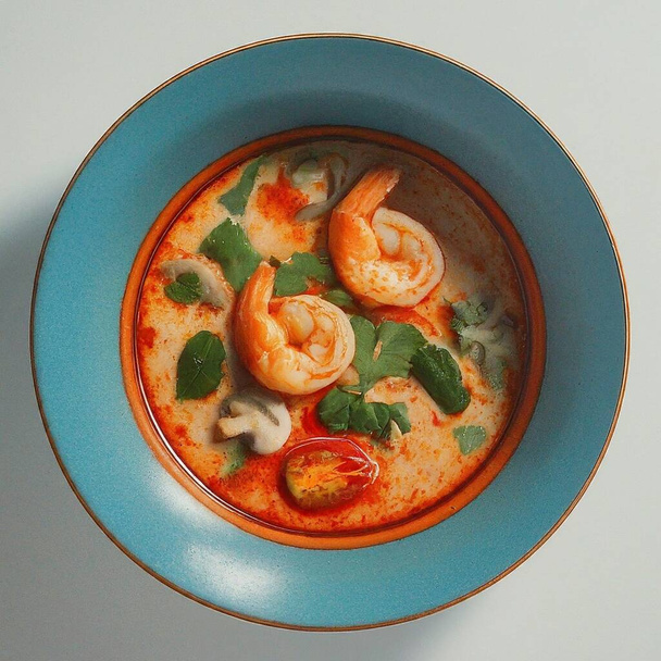 Egy tál fűszeres thai Tom Yum leves ül az asztalon, a gazdag vörös leves tele gyógynövényekkel, egy lédús garnélarák körettel, és egy darab mész ízesítésére. - Fotó, kép
