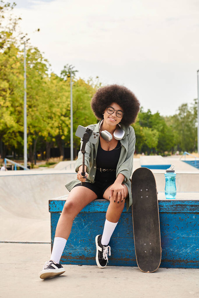 Uma jovem afro-americana com cabelo encaracolado senta-se em um parque de skate enquanto segura uma vara selfie - Foto, Imagem