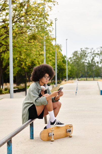 Młoda Afroamerykanka z kręconymi włosami siedzi na deskorolce, pochłonięta komórką w skateparku. - Zdjęcie, obraz