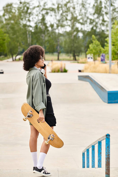 Mladá Afroameričanka s kudrnatými vlasy, drží skateboard při chatování na mobilním telefonu v pulzujícím venkovním skate parku. - Fotografie, Obrázek