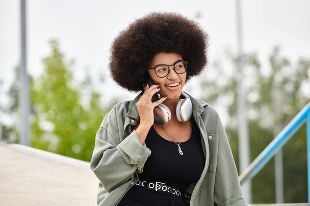 Μια κομψή γυναίκα με αφρο χτένισμα συνομιλίες στο smartphone της, ενώ απολαμβάνετε την ύπαιθρο. - Φωτογραφία, εικόνα