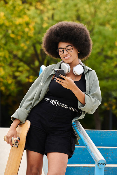 Femme afro-américaine énergique avec cheveux bouclés, écouteurs et planche à roulettes dans un skate park. - Photo, image