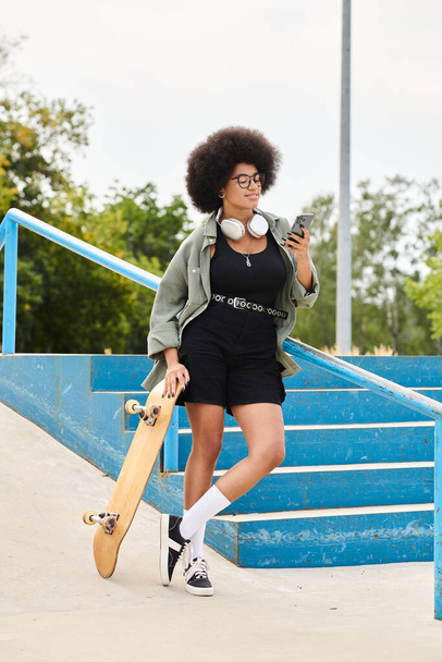 Een jonge Afro-Amerikaanse vrouw met krullend haar met een skateboard en een mobiele telefoon buiten in een skatepark. - Foto, afbeelding