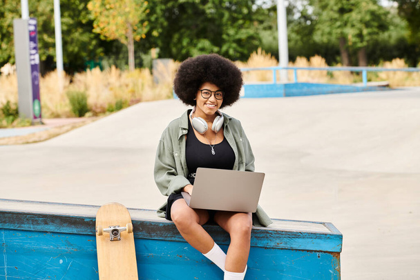 Una mujer negra con el pelo rizado se sienta en una caja azul usando un portátil. - Foto, imagen