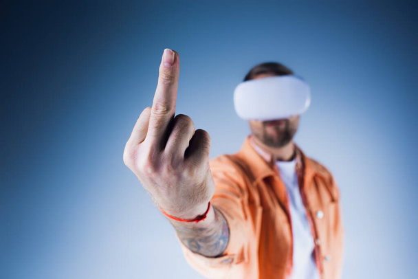 Mężczyzna, z zawiązanymi oczami i założonymi słuchawkami VR, pewnie wskazuje niewidzialny cel w studio, środkowy palec - Zdjęcie, obraz