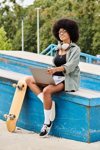 Een jonge Afro-Amerikaanse vrouw met krullend haar zittend op een bank met haar skateboard in een outdoor skate park. - Foto, afbeelding