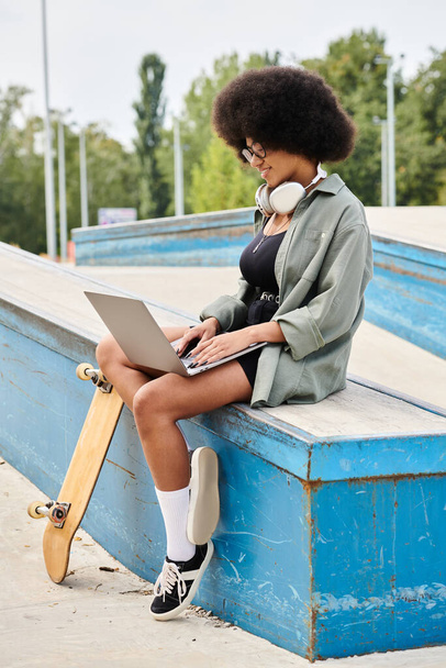 Eine junge Afroamerikanerin mit lockigem Haar sitzt auf einem Skateboard und tippt auf einem Laptop in einem Skatepark. - Foto, Bild