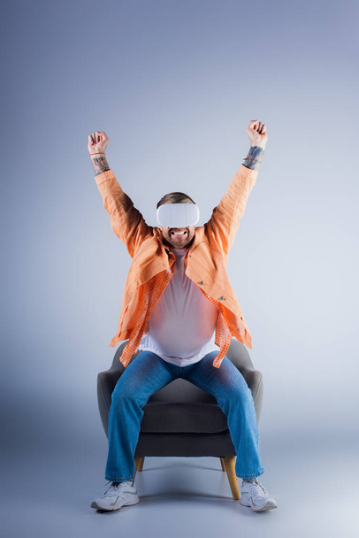Człowiek w zestawie słuchawkowym VR siedzi na krześle z ramionami podniesionymi w święto w studio. - Zdjęcie, obraz
