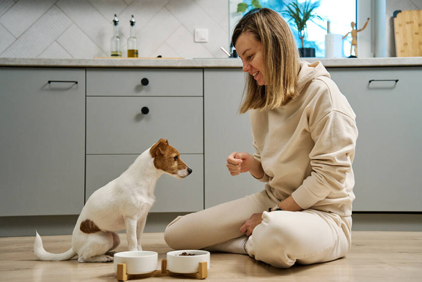 Kobieta umieszczenie miski żywności z paszą dla psa na podłodze w kuchni, Właścicielka spędza czas ze zwierzętami w domu, Karmienie zwierząt i opieki nad zwierzętami - Zdjęcie, obraz