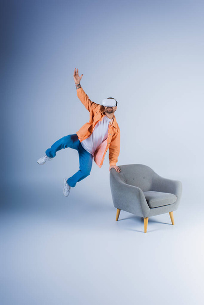 Una persona que lleva un auricular VR salta enérgicamente junto a una silla en un entorno de estudio. - Foto, imagen