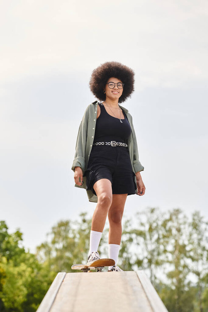 Μια νεαρή Αφρο-Αμερικανίδα με άφρο κάνει σκέιτμπορντ με χάρη σε μια ράμπα σε ένα υπαίθριο πάρκο για σκέιτ.. - Φωτογραφία, εικόνα