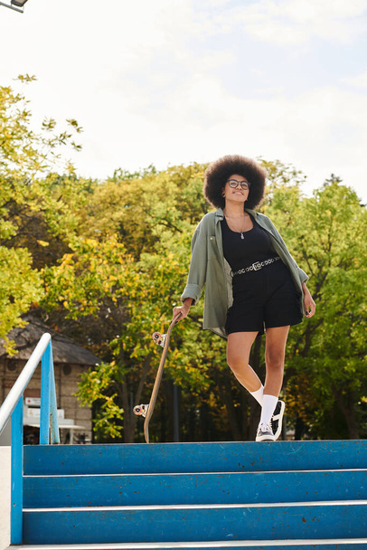 Uma jovem afro-americana com cabelo encaracolado sobe confiantemente um lance de escadas enquanto segura um skate. - Foto, Imagem