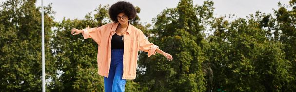 Młoda Afroamerykanka z kręconymi włosami z wdziękiem stoi na deskorolce, otoczona bujnymi drzewami w skateparku. - Zdjęcie, obraz