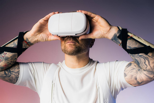 Un hombre con una camisa blanca sostiene un objeto blanco sobre su cabeza, inmerso en un auricular de realidad virtual en un entorno de estudio. - Foto, Imagen