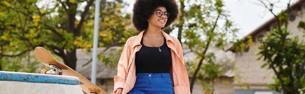Une jeune afro-américaine aux cheveux bouclés se tient à côté d'une planche à roulettes dans un parc, prête à frapper les rampes et à montrer ses compétences. - Photo, image