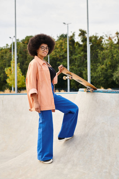 Młoda Afroamerykanka z kręconymi włosami umiejętnie stojąca na rampie na deskorolce w skateparku. - Zdjęcie, obraz