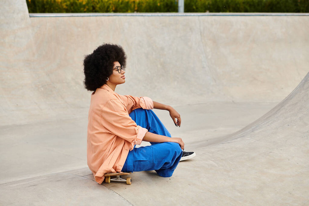 Mladá Afroameričanka s kudrnatými vlasy sebevědomě sedí na skateboardu v rušném skate parku, připravená plachtit. - Fotografie, Obrázek
