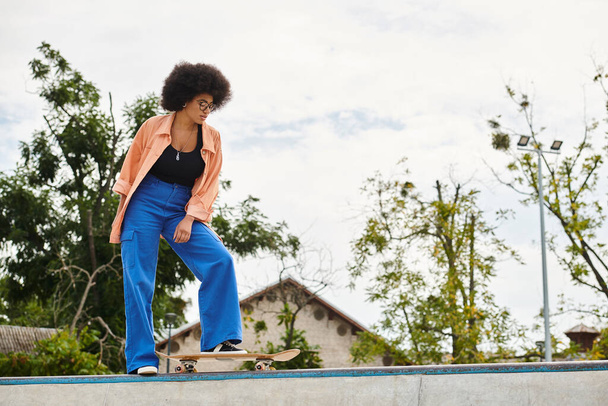 Young African American woman with curly hair riding skateboard on top of cement wall in outdoor skate park. - Photo, Image