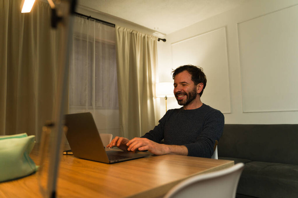 Opgewonden Kaukasische man glimlachend achter zijn bureau met de laptop en 's avonds vanuit huis werkend - Foto, afbeelding