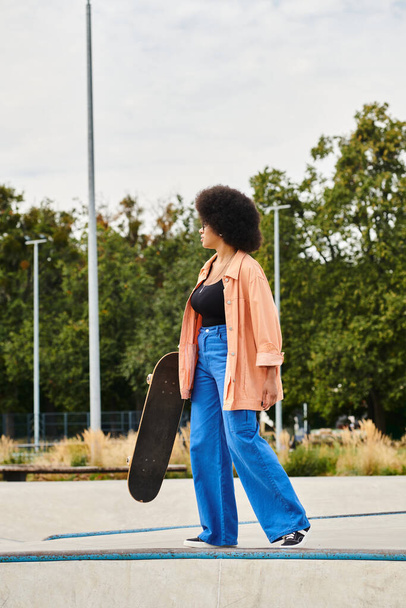 Νεαρή Αφρο-Αμερικανίδα γυναίκα με σγουρά μαλλιά περπατάει άνετα σε ένα πάρκο για σκέιτ, κρατώντας ένα σκέιτμπορντ στο χέρι της. - Φωτογραφία, εικόνα