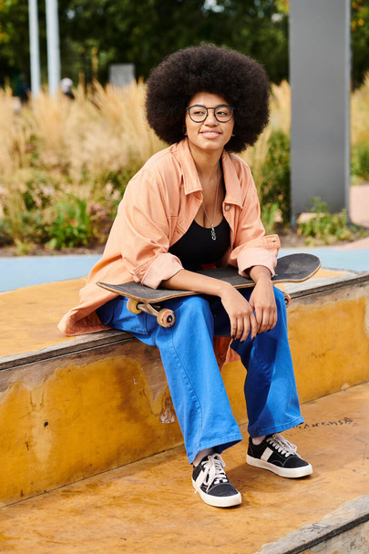 Una joven afroamericana con el pelo rizado se sienta en una cornisa con su monopatín en un vibrante parque de skate al aire libre. - Foto, imagen