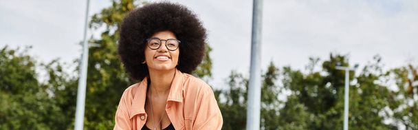 Tyylikäs nuori afroamerikkalainen nainen, jolla on kiharat hiukset silmälasit ja vaaleanpunainen paita rullalautailu ulkona luistelupuistossa. - Valokuva, kuva
