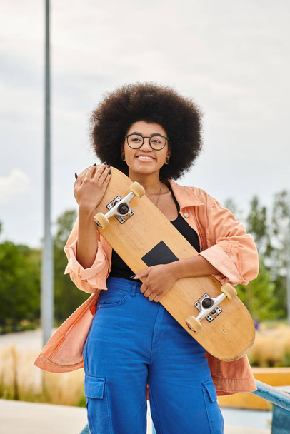 Stylowa Afroamerykanka z afro hairdo pewnie trzyma deskorolkę w skateparku. - Zdjęcie, obraz
