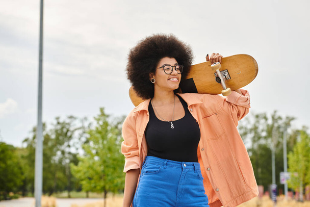 Egy fiatal, göndör hajú afro-amerikai nő gördeszkát tart az arcához egy gördeszkás parkban.. - Fotó, kép
