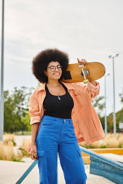 Μια νεαρή Αφρο-Αμερικανίδα γυναίκα με αφρο με αυτοπεποίθηση κρατά ένα skateboard σε ένα ζωντανό πάρκο skate. - Φωτογραφία, εικόνα