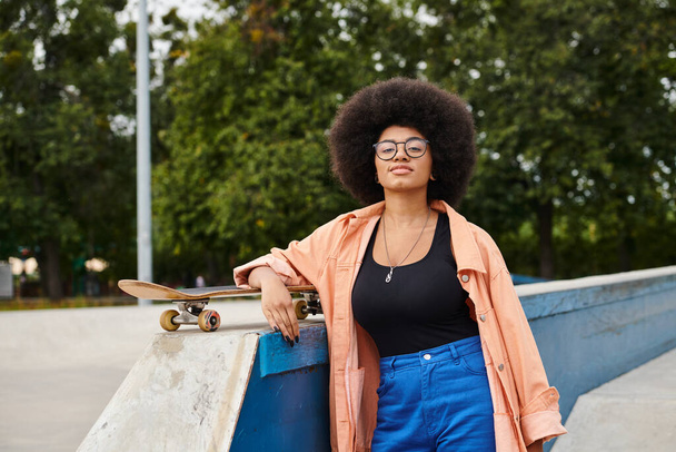 Een jonge Afro-Amerikaanse vrouw met krullend haar staat vol vertrouwen naast een skateboard op een skateparkhelling. - Foto, afbeelding