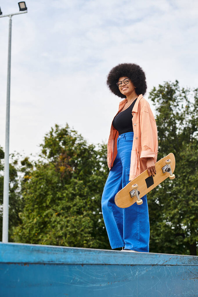 Молодая афроамериканка с вьющимися волосами уверенно держит скейтборд, стоящий на рампе в скейт-парке. - Фото, изображение