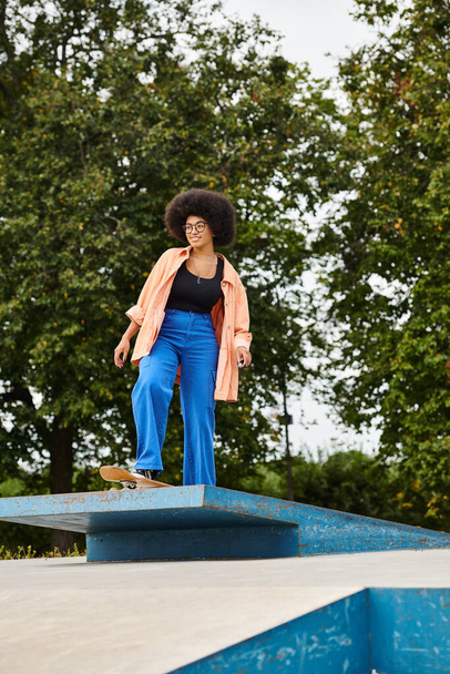 Eine junge Afroamerikanerin mit lockigem Haar steht anmutig auf einem blauen Gegenstand in einem Skatepark. - Foto, Bild