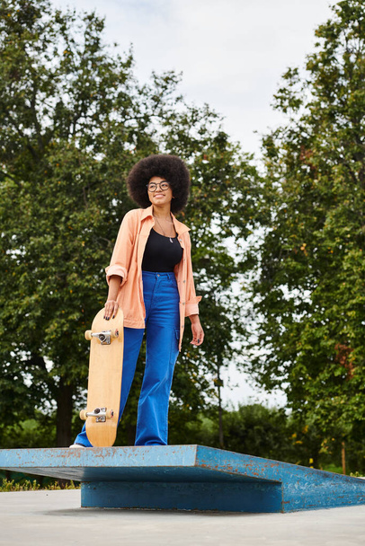 Junge Afroamerikanerin mit lockigem Haar hält ein Skateboard auf einer blauen Plattform in einem Skatepark. - Foto, Bild
