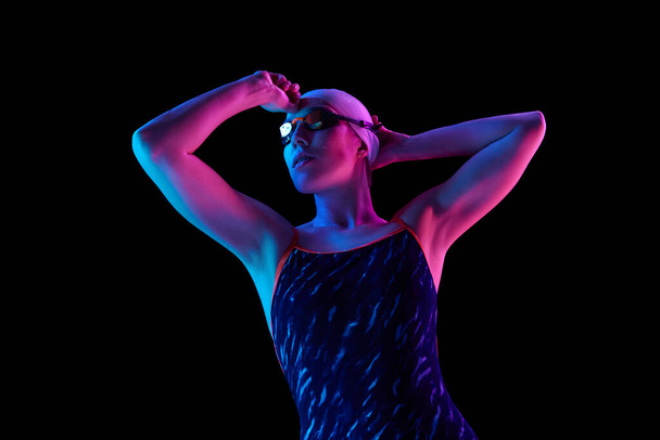 Confiant, concentré sur la compétition jeune nageur en uniforme de natation posant dans une lumière au néon vibrante sur fond de studio noir. Concept de sport professionnel, mouvement, force et puissance. Publicité - Photo, image