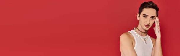 svůdný gay muž v elegantním oblečení s doplňky na červeném pozadí a při pohledu na fotoaparát, banner - Fotografie, Obrázek