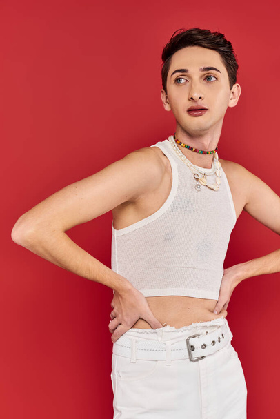 молодой модный гей в повседневной одежде с аксессуарами позируя на красном фоне и глядя в сторону - Фото, изображение