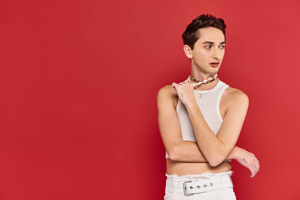 attractive fashionable gay man in casual attire with accessories on red backdrop and looking away - Foto, immagini