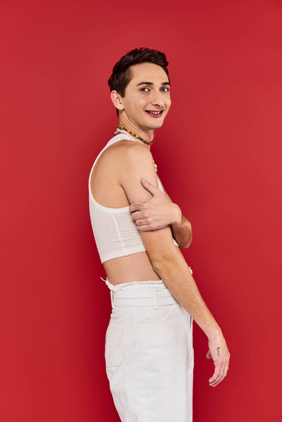 alegre guapo gay hombre con elegante accesorios en blanco atuendo mirando la cámara en rojo telón de fondo - Foto, Imagen