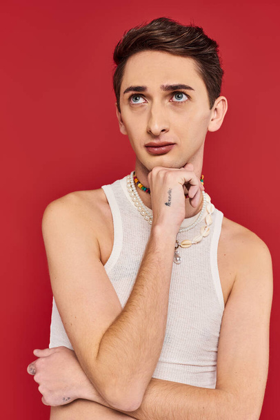 handsome fashionable gay man in casual attire with accessories on red backdrop and looking away - Zdjęcie, obraz