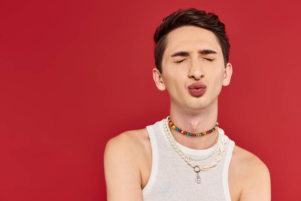 appealing young gay man in stylish white attire with closed eyes and pounded lips on red backdrop - 写真・画像